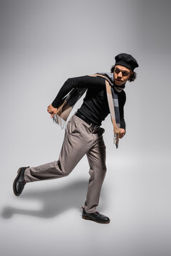 full length of african american man in black beret and sweater with trousers running and looking away on grey background
