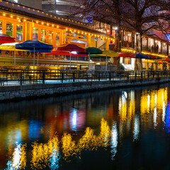 Vibrant colors of boardwalk umbrellas and water reflections on San Antonio River Walk canal on a quiet winter night with empty seats in Texas, USA