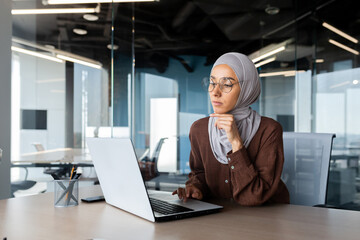 Successful businesswoman thinking at workplace, muslim woman in hijab using laptop at work, serious...