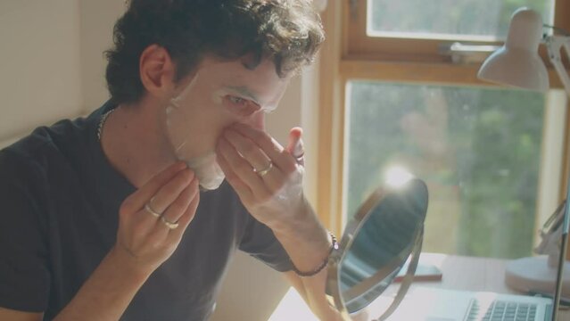 Young man looking at his reflection in tabletop mirror while applying moisturizing sheet mask to face in morning at home