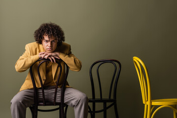 brunette african american man in trendy casual outfit sitting on black chair and looking at camera on olive grey background
