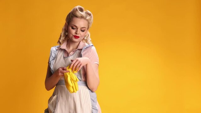 Beautiful stylish girl, housewife in retro apron, putting on rubber gloves for cleaning over yellow studio background. Concept of retro fashion, beauty, attraction, 50s, 60s. Pin-up style. Vintage