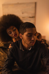 curly african american woman touching shoulders of man in black shirt sitting in bedroom at night