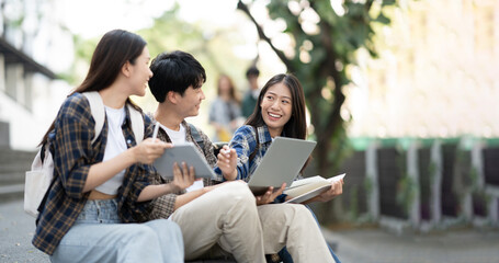 Young Asian college students reading books, studying on laptop, preparing for exam or working on group project while sitting on staircase of college campus.