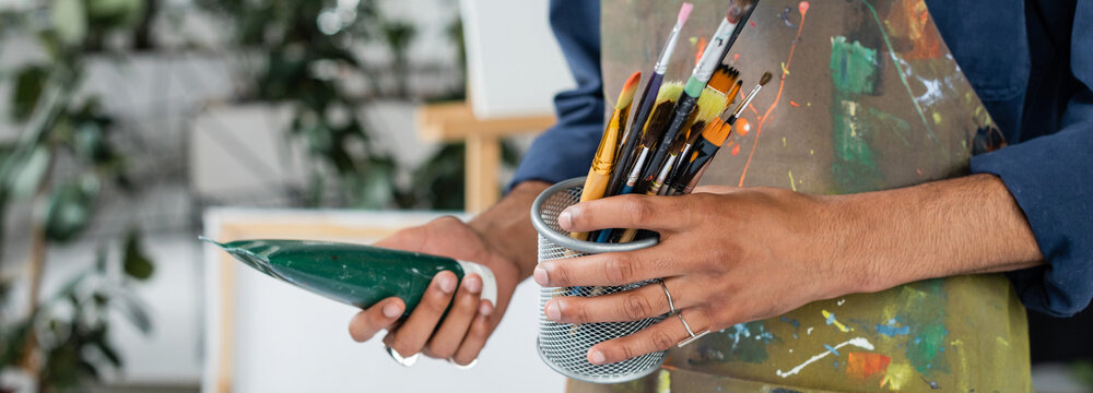 Cropped view of afrcan american artist in apron holding paintbrushes and paint, banner.