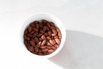 Selective focus flat lay side lit view of dry-roasted smoked almonds in lightly speckled white bowl on ceramic background