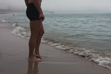 A girl's feet on the ocean in the fog. A girl stands on a sandy beach by the water in cloudy weather.