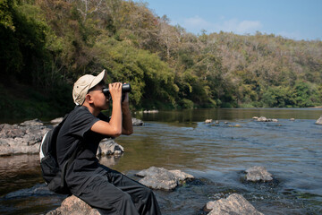 Asian boy wearing black t-shirt holding a binoculars sitting on stone by the river, looking through lens to see birds on tree branches and on sky in local national park during his summer vacation.