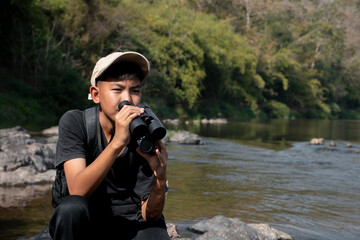 Asian boys wearing black t-shirt holding a binoculars sitting on stone by the river flowing down from the mountains in national park to observe fish in the river and birds on tree branches and on sky.