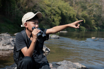 Asian boy wearing black t-shirt holding a binoculars sitting on stone by the river, pointing to birds on trees  in national park to observe fish in the river and birds on tree branches and on sky.