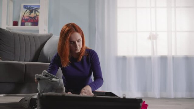 Redhead woman takes clothes out of open black suitcase