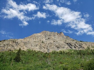 Outdoor daytime nature landscape image looking up from below at a mountain peak that has green grass and forest at the base and blue sky with fluffy white clouds above.