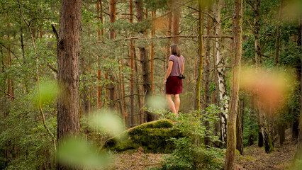 Fototapeta na wymiar A young woman in a red skirt is stand on a rock in a forest. In the bokeh foreground are some blured leaves.