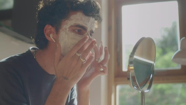 Medium close up shot of young man in wireless earbuds listening to music while applying clay mask to face in front of tabletop mirror at home