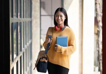 Young Asian Student Standing at Entrance to University Building. Getting rady to go to classroom holding books and bag. - obrazy, fototapety, plakaty