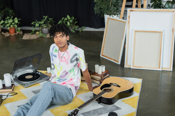 African american artist looking at camera near acoustic guitar and canvases in workshop.
