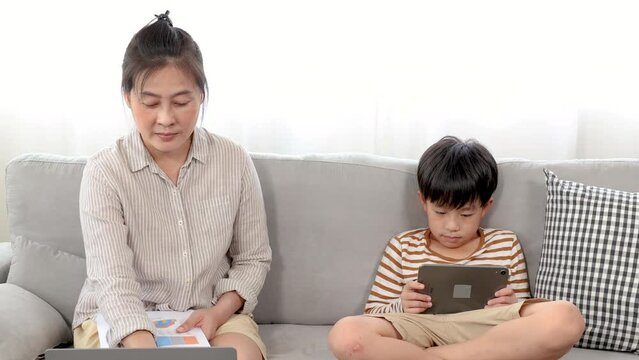 Asian single mother sitting on sofa using laptop working from home and handsome son play game, watching movie with tablet next to mom. Family using different electronic devices. enjoy social.
