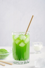 Cold refreshing healthy matcha made of green tea leaves ground powder served with ice and drinking paper straw on white wooden kitchen table with ingredients as organic antioxidant vitamin drink