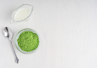 Top view of organic detox matcha ground powder of green tea leaves served in glass bowl with plant milk and spoon on white wooden background with copy space used as ingredient of healthy latte drink