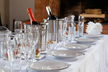 Exquisitely set banquet table with white plates, wine glasses and a buckets of champagne indoor next to the fireplace, selective focus.