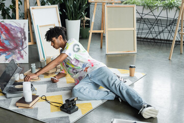 Side view of african american artist holding smartphone and using record player near books and vintage camera in studio.