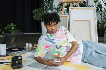 African american artist using smartphone near vintage camera and blurred canvases.
