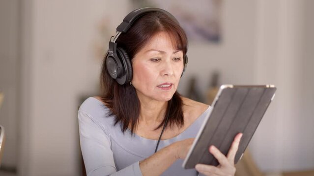 Stylish attractive woman wearing headphones relaxes listening to music reading mobile tablet story sitting in comfortable home chair