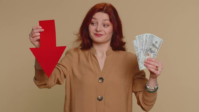 Upset redhead woman showing red arrow pointing down, concept of downgrade, unsuccessful business, fall of stock market money dollar exchange rate bankruptcy fail. Young ginger girl on beige background