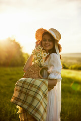 a beautiful woman in a hat and a light dress walks through a chamomile field with a plaid in her hands