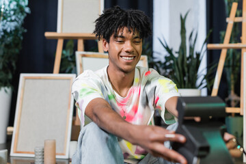 Smiling african american artist taking photo on blurred vintage camera in studio.