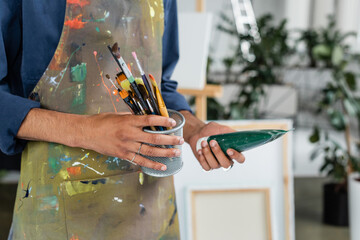 Cropped view of african american artist in apron holding paintbrushes and paint tube.