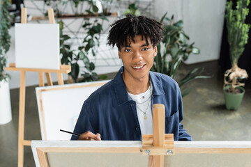 Smiling african american artist holding paintbrush and looking at camera near canvas in studio.