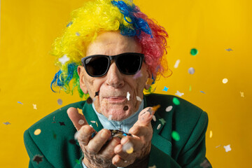 Front view of an old man dressed for carnival and blowing confetti with sunglasses, green jacket, colored wig