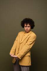 Fototapeta na wymiar trendy african american man in yellow blazer looking at camera while posing with clenched hands on olive grey background