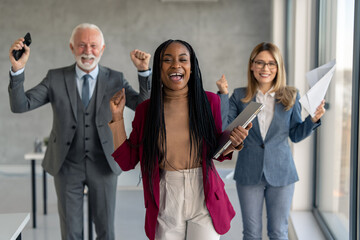 Beautiful confident smiling African-American businesswoman team leader standing in front of her successful team, holding laptop and cheering with fists up. Successful diverse business team in office.