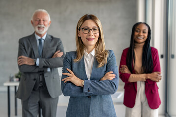 Shot of beautiful confident smiling businesswoman in suit wearing glasses with arms crossed standing in front of her successful team. Thriving company culture concept.