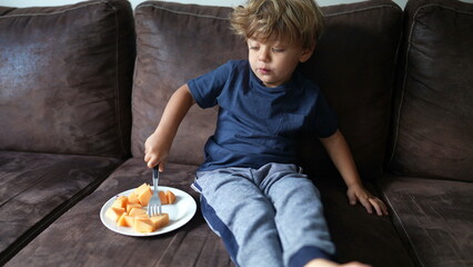 Adorable child eating melon fruit sitting on couch small boy eats healthy snack fruit with fork