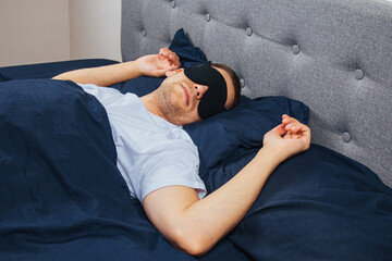 A man wrapped in a blanket wearing a sleep mask on a gray background. Sleeping at home in his bed....