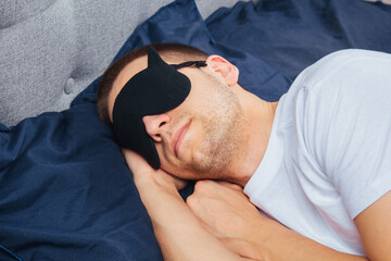 Side view of a young smiling cheerful man in a sleep mask in pajamas resting, relaxing at home, wrapping in a blanket, walking isolated on a dark blue background. 