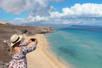  Mujer rubia con vestido floreado con sombrero de paja tomando una foto con su teléfono móvil de la playa de arena blanca y el mar turquesa de Jandia en Fuerteventura, Islas canarias.
