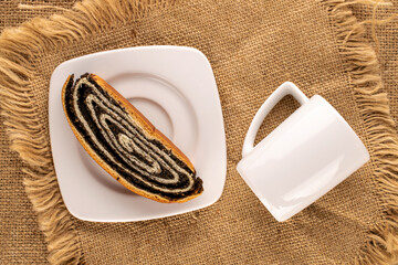 Fototapeta na wymiar One piece of appetizing poppy seed roll on white ceramic plate with cup on jute cloth, macro, top view.
