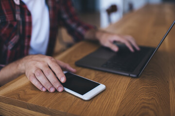 Cropped view of male programmer using digital laptop computer and smartphone gadget while doing...