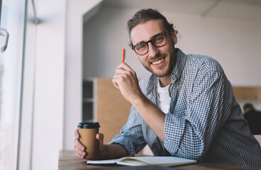 Portrait of cheerful male in optical spectacles smiling at camera during coffee break and time for studying in cafeteria, happy Caucaisan student with education textbook and takeaway cup posing