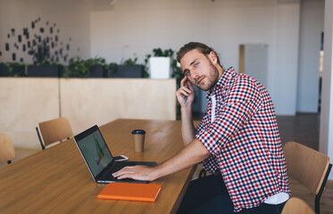 Portrait of skilled Caucaisan IT professional looking at camera during distance job in coworking space, millennial man in smart casual wear posing at table desktop with modern laptop computer