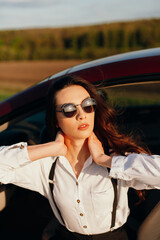 young beautiful woman in a white shirt and sunglasses sitting in a red car with a white interior and with open doors. sunny day. 
red car in the field. the wind blows the girl's hair.