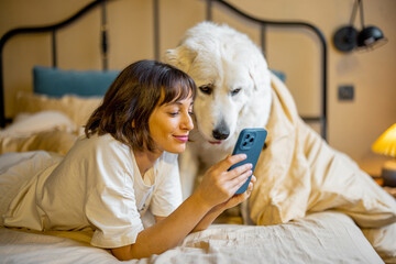 Young woman uses smart phone while lying with her cute adorable dog in bed at cozy bedroom in beige tones. Concept of leisure time with pets - 567833022