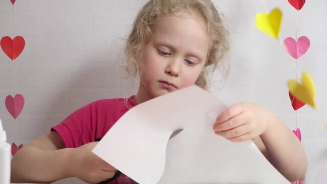 a girl of 7 years old cuts out a heart from colored paper sitting at the table