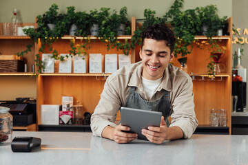 Smiling coffee shop owner at the counter with digital tablet. Male bartender in an apron leaning...