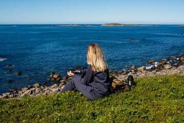 Fototapeta premium Woman with a cup of hot tea and a thermos sits on the ocean and admires the picturesque landscape. Beautiful nature. Harmony, relaxed lifestyle. Travel, adventure. Explore Northern Norway. Scandinavia