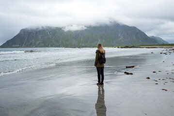 Blond hair girl with a backpack walks along an empty sandy ocean beach. Town and mountains. Beautiful nature landscape. Lonely girl. Wanderlust. Travel, adventure, lifestyle. Explore North, Norway 
